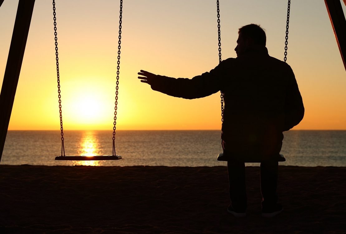 Homem sentando em um balanço estendendo a sua mão para o lado esquerdo ao encontro do outro banco (do qual não tem nenhuma pessoa) onde o brilho do nascer do sol aparece.